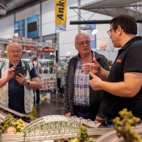Zwei Männer unterhalten sich über Modelleisenbahnen. Ein weiterer Mann hält ein Smartphone in der Hand.