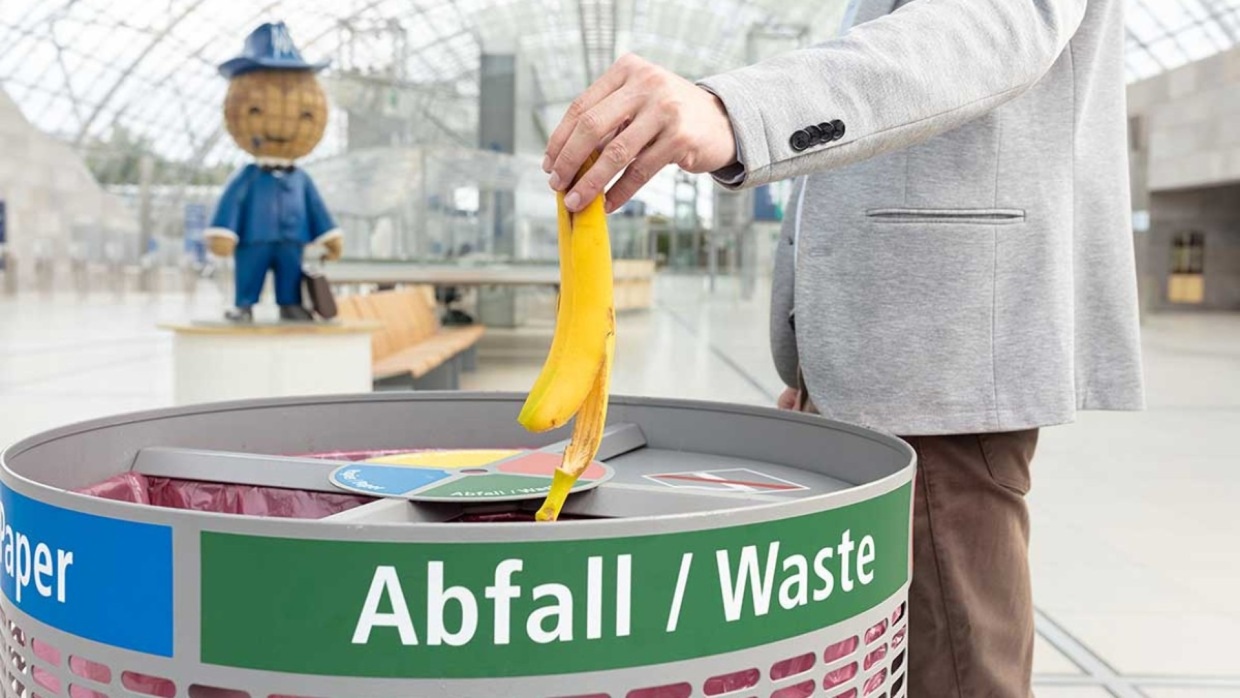 Person wirft eine Bananenschale in einen Abfallbehälter mit Sortierfächern in der Glashalle. Im Hintergrund ist das Messemännchen zu sehen.