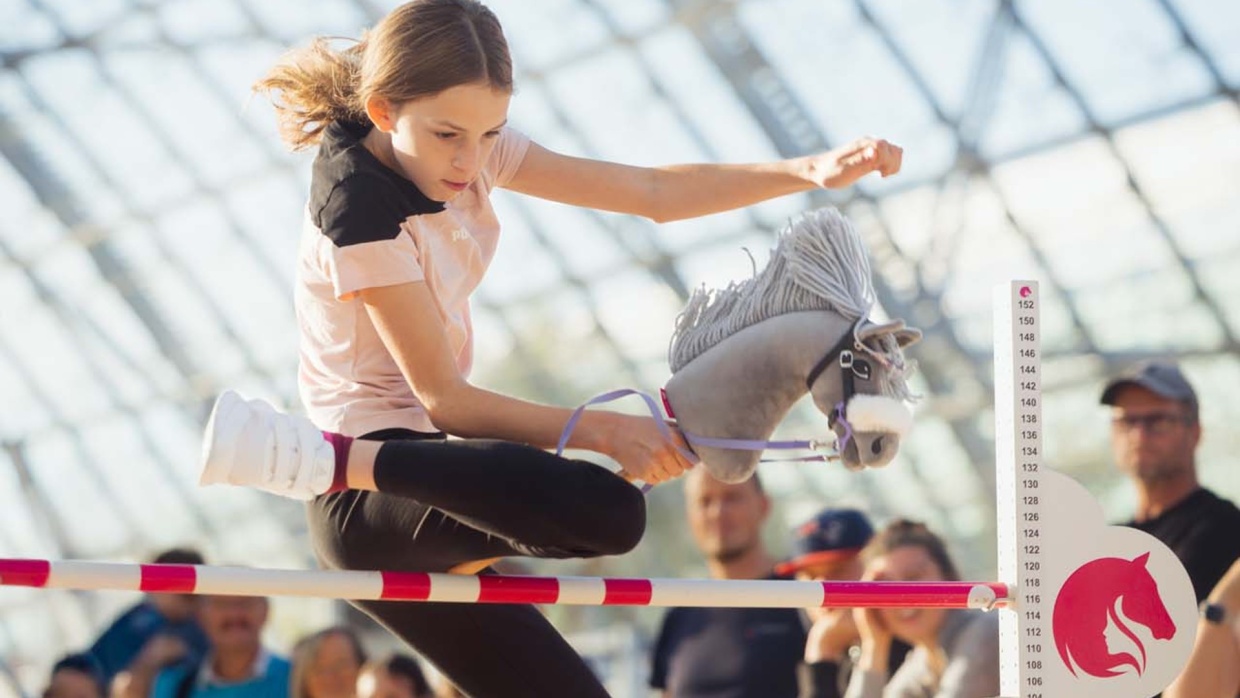 Person springt bei einem Hobby Horsing Turnier auf der Hobbymesse Leipzig 2025 über ein Hindernis. Im Hintergrund schauen Begeistert Hobby-Horser dem Sprungwettbewerb zu.
