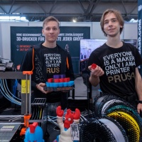 Zwei Männer stehen hinter einem 3D-Drucker und zeigen in die Kamera was sie gedruckt haben.