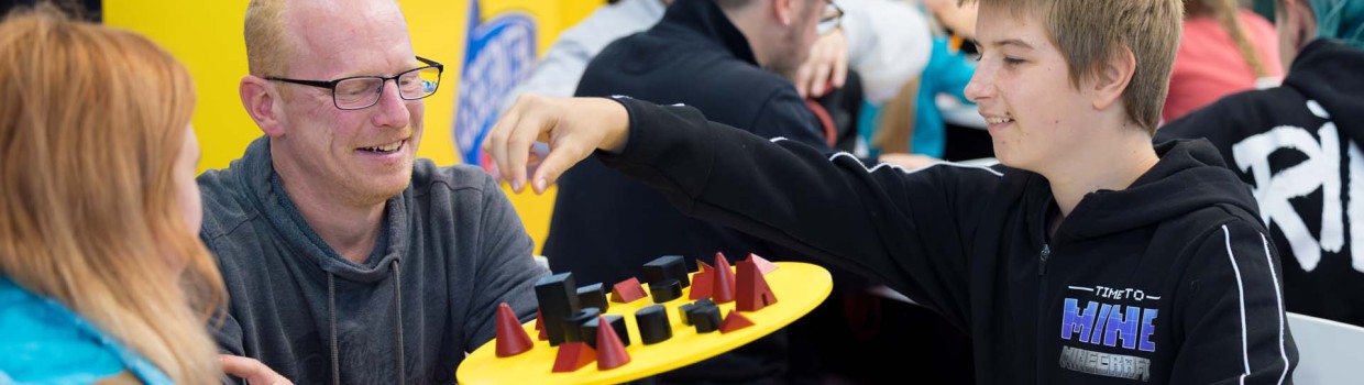 Drei Personen spielen gemeinschaftlich ein Geschicklichkeitsspiel auf der Hobbymesse Leipzig 2025. Im Hintergrund stehen mehr Spieletisch, wo Menschen Brettspiele ausprobieren.