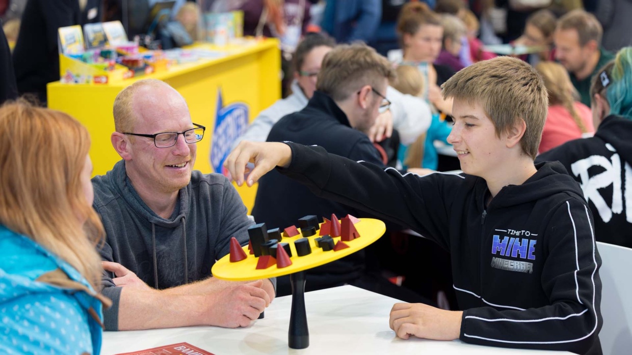 Drei Personen spielen gemeinschaftlich ein Geschicklichkeitsspiel auf der Hobbymesse Leipzig 2025. Im Hintergrund stehen mehr Spieletisch, wo Menschen Brettspiele ausprobieren.