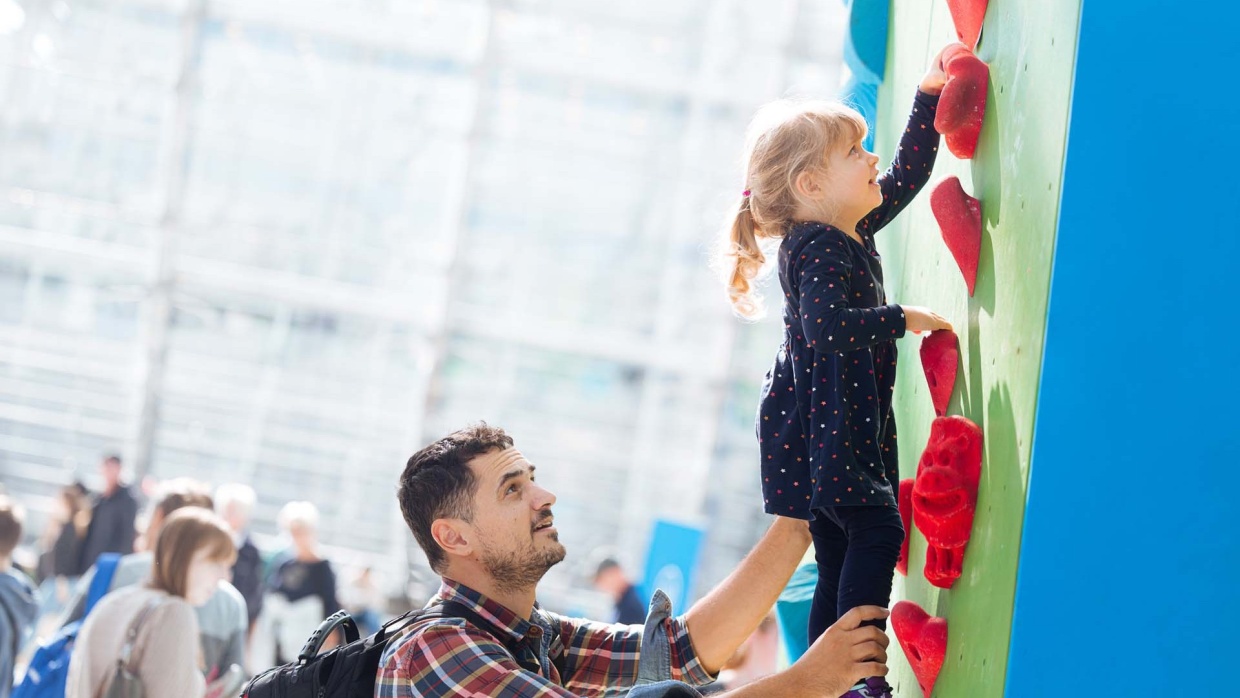 Eine Person hilft ihrem Kind auf der Hobbymesse Leipzig 2025 beim Klettern beziehungsweise Bouldern in der Glashalle der Leipziger Messe am Kletterturm.